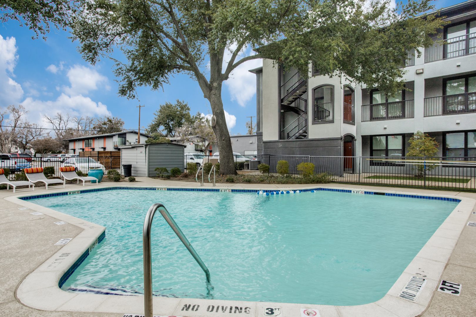 A bright outdoor pool courtyard invites relaxation and leisurely swimming in clear, inviting water. The scene centers on the pool's edge and ladder as the primary features for a refreshing lifestyle. Rounded concrete coping frames the blue-tiled pool, with safety depth markings along the deck and a stainless-steel handrail for easy access. Nearby, patio lounge chairs and umbrellas rest along the fence line, suggesting a comfortable spot to unwind between dips. Mature trees and well-kept landscaping create a calm, sunny atmosphere around the modern multi-story building with interior stair access and balconies.