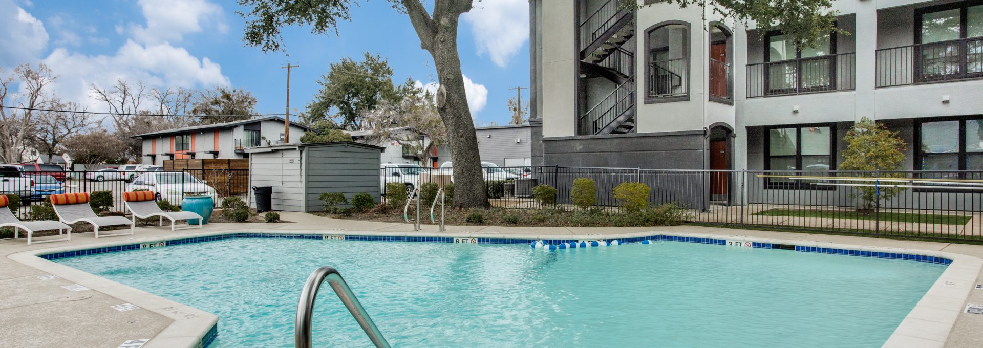 A bright outdoor pool courtyard invites relaxation and leisurely swimming in clear, inviting water. The scene centers on the pool’s edge and ladder as the primary features for a refreshing lifestyle. Rounded concrete coping frames the blue-tiled pool, with safety depth markings along the deck and a stainless-steel handrail for easy access. Nearby, patio lounge chairs and umbrellas rest along the fence line, suggesting a comfortable spot to unwind between dips. Mature trees and well-kept landscaping create a calm, sunny atmosphere around the modern multi-story building with interior stair access and balconies.
