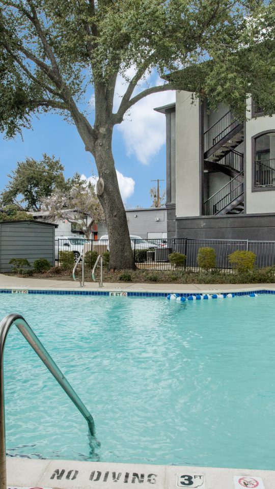 A bright outdoor pool courtyard invites relaxation and leisurely swimming in clear, inviting water. The scene centers on the pool’s edge and ladder as the primary features for a refreshing lifestyle. Rounded concrete coping frames the blue-tiled pool, with safety depth markings along the deck and a stainless-steel handrail for easy access. Nearby, patio lounge chairs and umbrellas rest along the fence line, suggesting a comfortable spot to unwind between dips. Mature trees and well-kept landscaping create a calm, sunny atmosphere around the modern multi-story building with interior stair access and balconies.