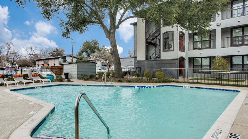 A bright outdoor pool courtyard invites relaxation and leisurely swimming in clear, inviting water. The scene centers on the pool&acirc;s edge and ladder as the primary features for a refreshing lifestyle. Rounded concrete coping frames the blue-tiled pool, with safety depth markings along the deck and a stainless-steel handrail for easy access. Nearby, patio lounge chairs and umbrellas rest along the fence line, suggesting a comfortable spot to unwind between dips. Mature trees and well-kept landscaping create a calm, sunny atmosphere around the modern multi-story building with interior stair access and balconies.