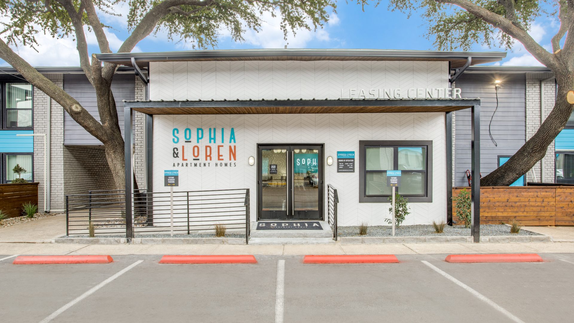 A quiet, inviting apartment community entrance greets visitors under a wide canopy. The primary focus is the main doorway and signage for SOPHIA & LOREN, highlighting a welcoming lifestyle and easy arrival. The modern facade features clean white brick with geometric wall panels, framed entry doors, and glass sidelights that let in light. A sturdy metal railing borders the steps, while landscaped gravel beds and small plants soften the walkway. Expansive trees arch overhead, enhancing the resort-like, well-kept feel of the building exterior.