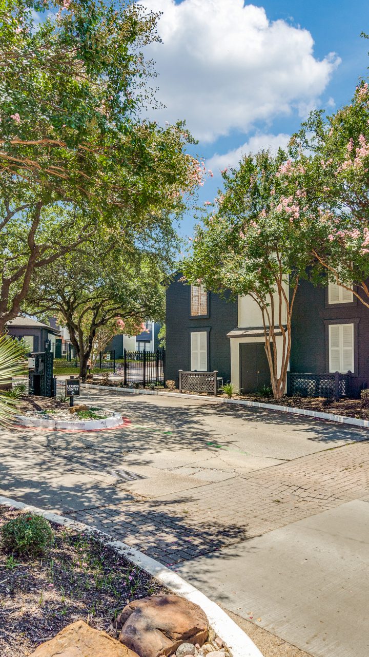 Landscaped entrance with mature trees and apartment buildings at Evaline Apartments in North Dallas, Texas