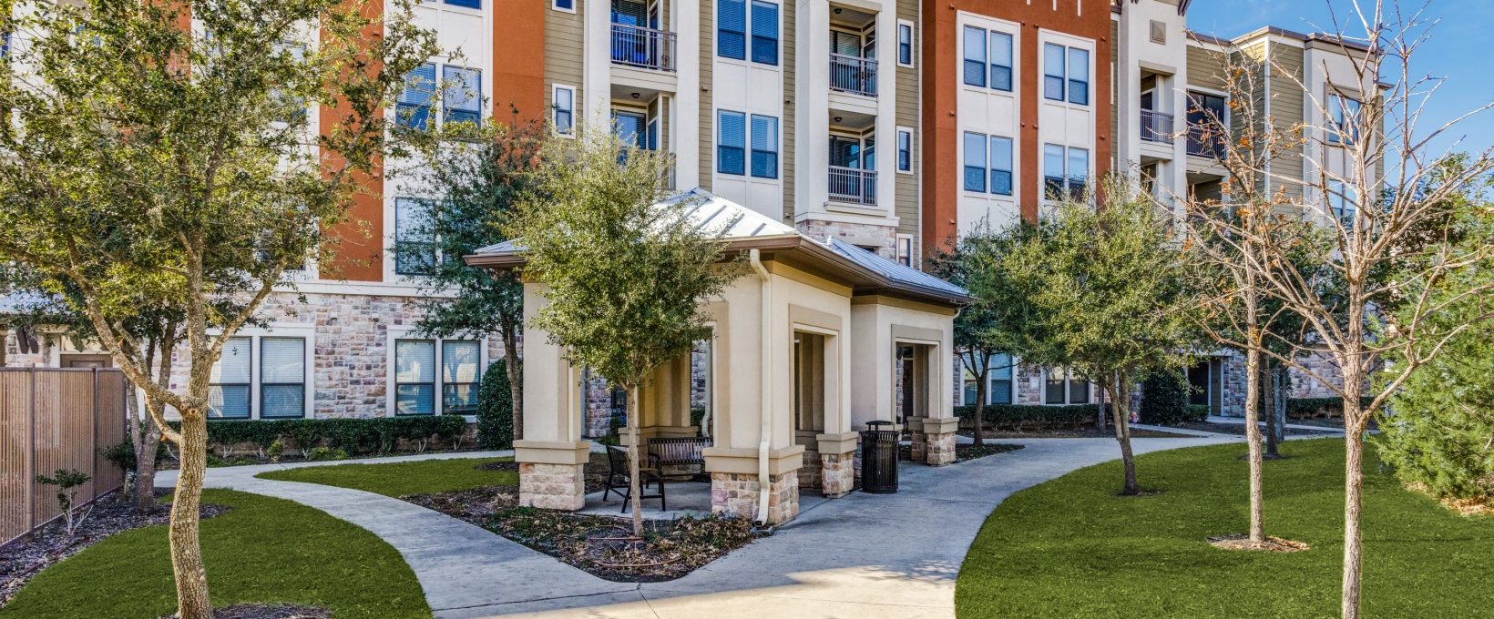 the front of an apartment complex with landscaping and trees at The  Carson @ Twin Creeks