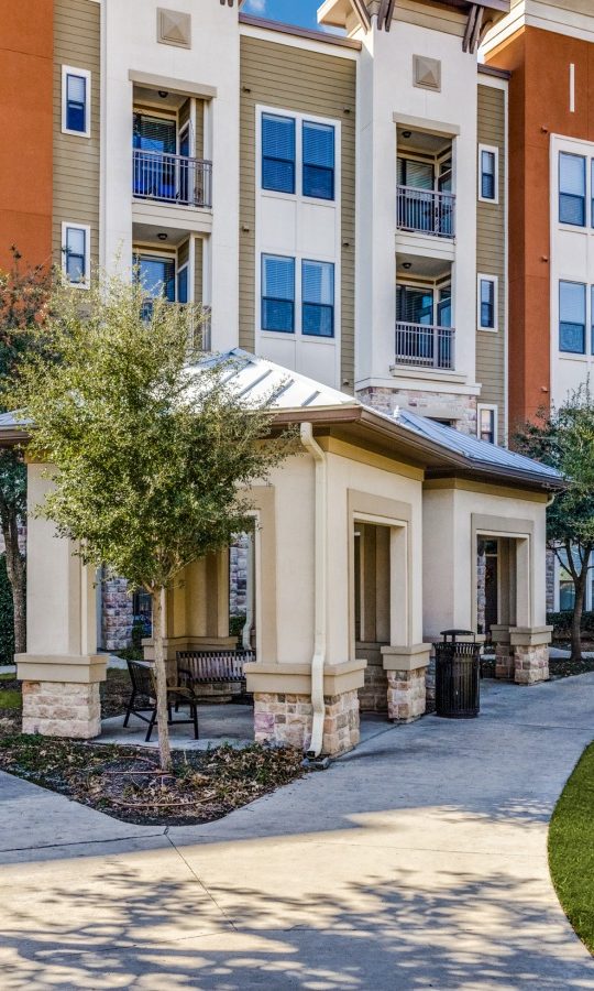 the front of an apartment complex with landscaping and trees at The  Carson @ Twin Creeks