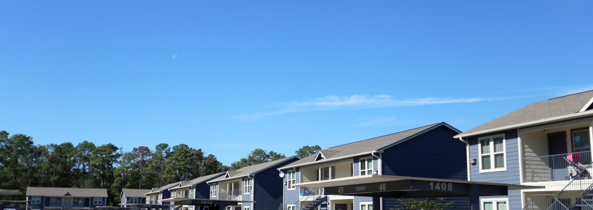 A serene apartment complex on a bright, sunny day with multiple buildings featuring blue and beige exteriors. The parking lot is filled with various cars, indicating the presence of residents or visitors. The buildings have covered entrances and balconies, suggesting comfortable and convenient living spaces. The area is surrounded by lush green trees, contributing to a peaceful and inviting atmosphere.